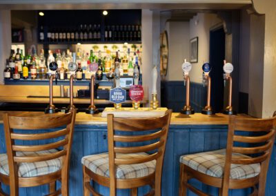 Picture of the bar with a large number of bottle in the background, wooden bar table and three tall chairs.